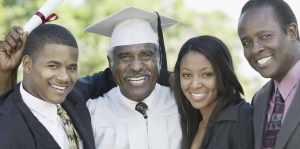 A joyful group photo of four people, including an older man wearing a white graduation cap and gown, holding a diploma. He is smiling broadly, surrounded by three younger adults — two men in suits and a woman in business attire — all smiling and posing closely together. The background features soft-focus greenery, suggesting the picture was taken outdoors on a sunny day.